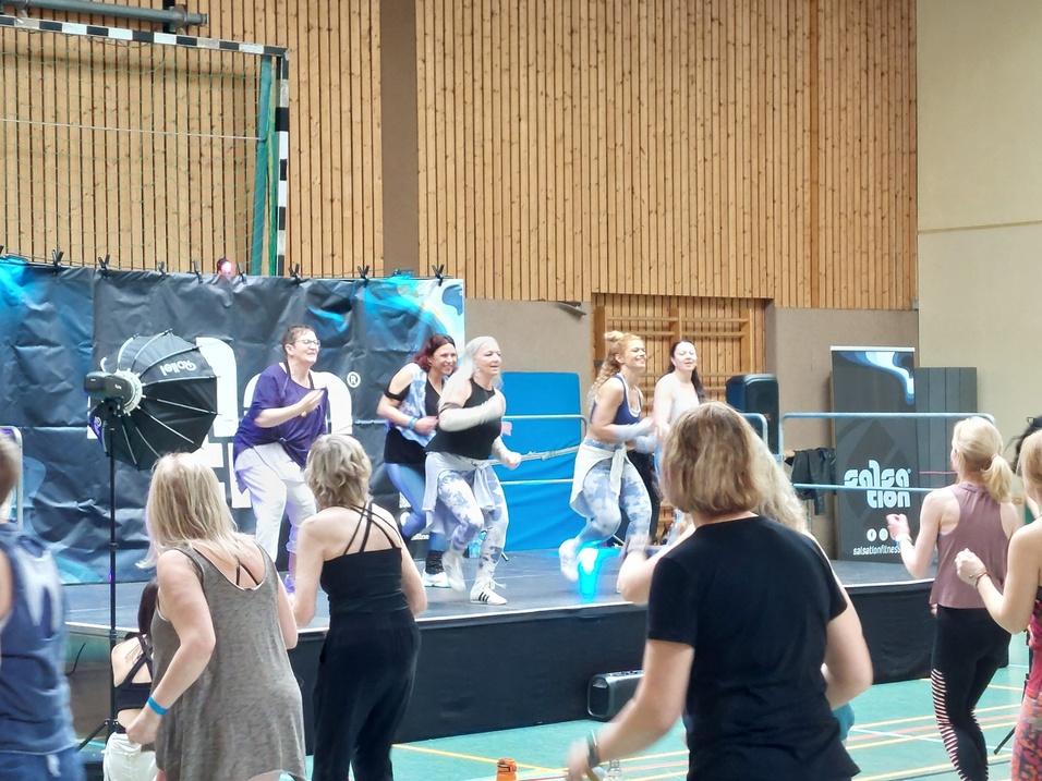 Gruppen von Frauen tanzen auf einer Bühne in einer Sporthalle, umgeben von Musik-Equipment und einem großen Banner.