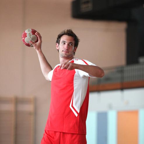 Junger Handballspieler in rotem Trikot wirft einen Handball in einer Sporthalle.
