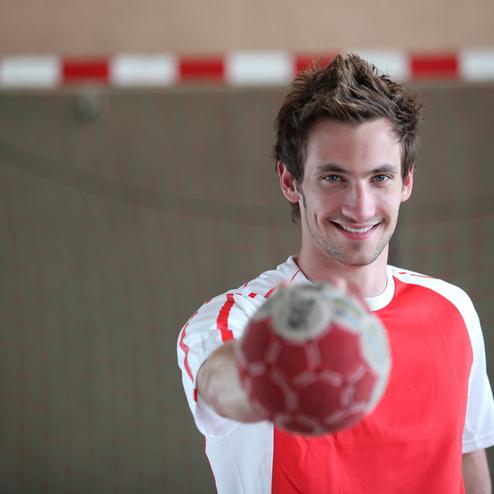 L&auml;chelnder Handballspieler h&auml;lt einen Handball in Richtung Kamera, im Hintergrund ein Handballtor.