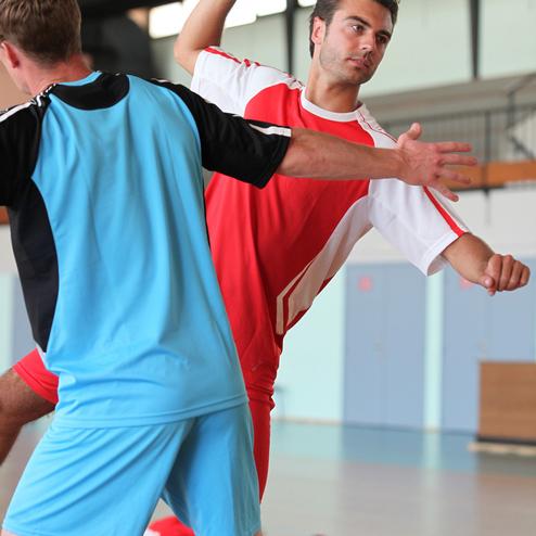 Spieler in rot wirft einen Handball, w&auml;hrend ein gegnerischer Spieler in blau ihn blockiert. Sporthalle im Hintergrund.
