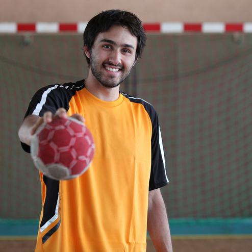 Junger Mann in orange-schwarzem Trikot h&auml;lt einen Handball vor einem Handballtor in einer Sporthalle.