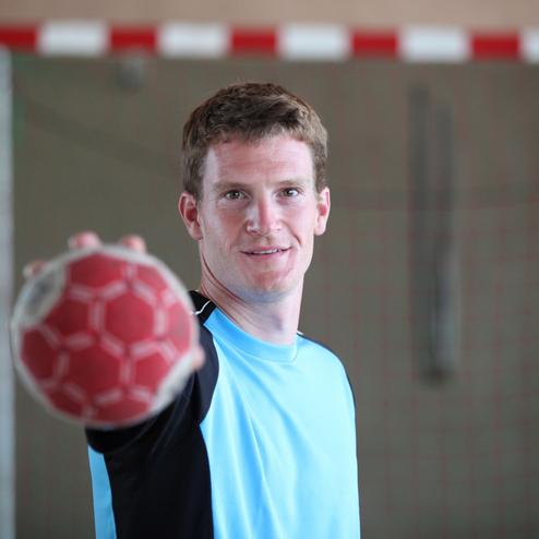 Mann in blauer Sportbekleidung h&auml;lt einen Handball mit einer Hand vor einem Handballtor.