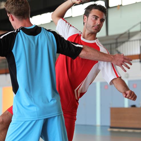 Zwei Handballspieler in Aktion, einer wirft den Ball, w&auml;hrend der andere ihn verteidigt. Sporthalle im Hintergrund.