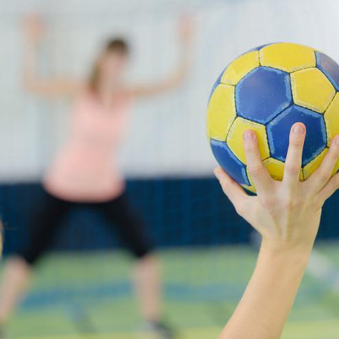 Hand mit gelb-blauem Handball im Vordergrund, im Hintergrund steht eine Spielerin vor dem Tor.