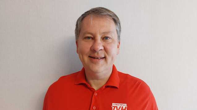 Mann mit kurzem, grauen Haar trägt ein rotes Poloshirt mit Logo vor hellem Hintergrund.