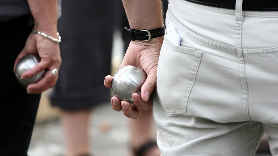 Zwei Personen halten silberne Kugeln in der Hand, während sie auf einem Platz an einem Boule-Spiel teilnehmen.