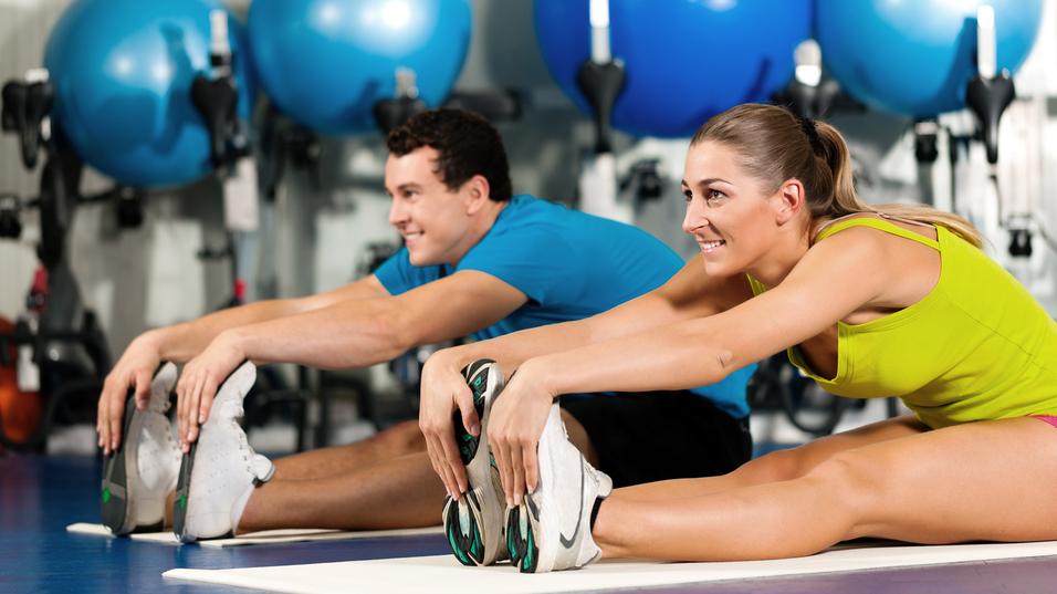 Zwei Personen dehnen auf Matten in einem Fitnessstudio, bunte Gymnastikbälle im Hintergrund.