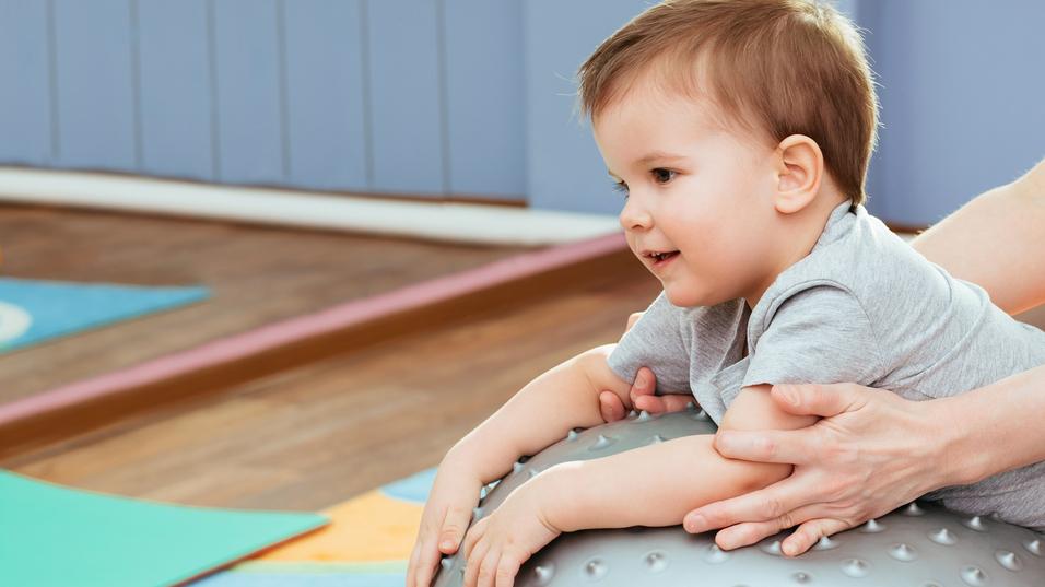 Kleines Kind auf einer großen, grauen Gymnastikball, während eine Hand es sanft stützt, in einem Spielraum.