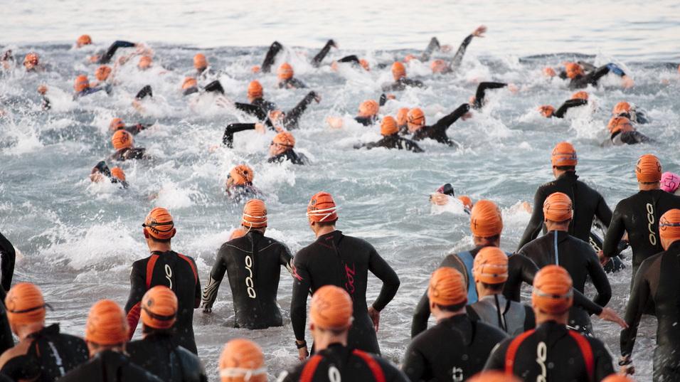 Gruppe von Schwimmern in Neoprenanzügen mit orangefarbenen Badekappen, die ins Wasser starten.
